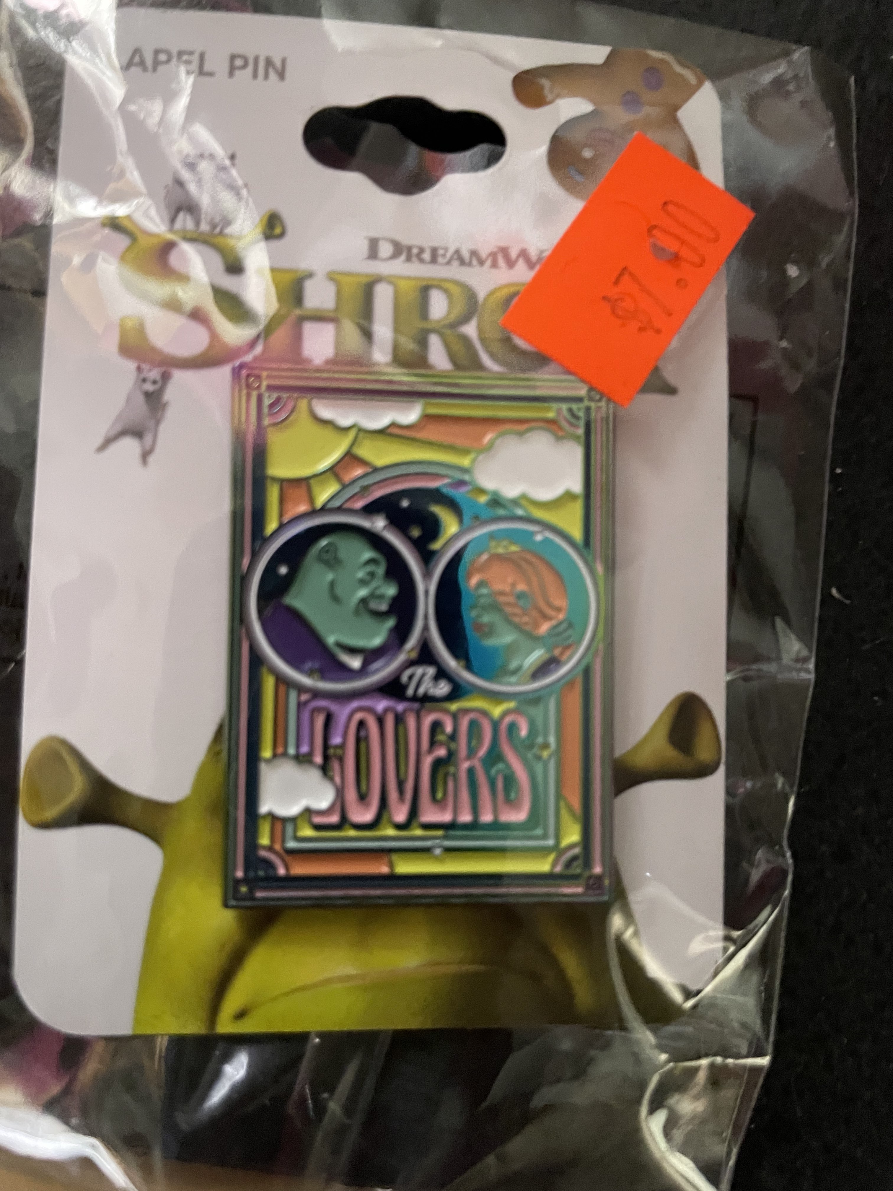 A LAPEL PIN OF SHREK AND FIONA FROM SHREK FACING EACHOTHER. BELOW THEM IT READS 'THE LOVERS'.